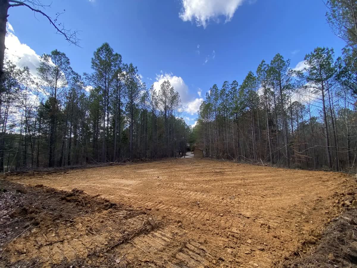 A building pad cleared and graded to red clay, tall pines on both sides.